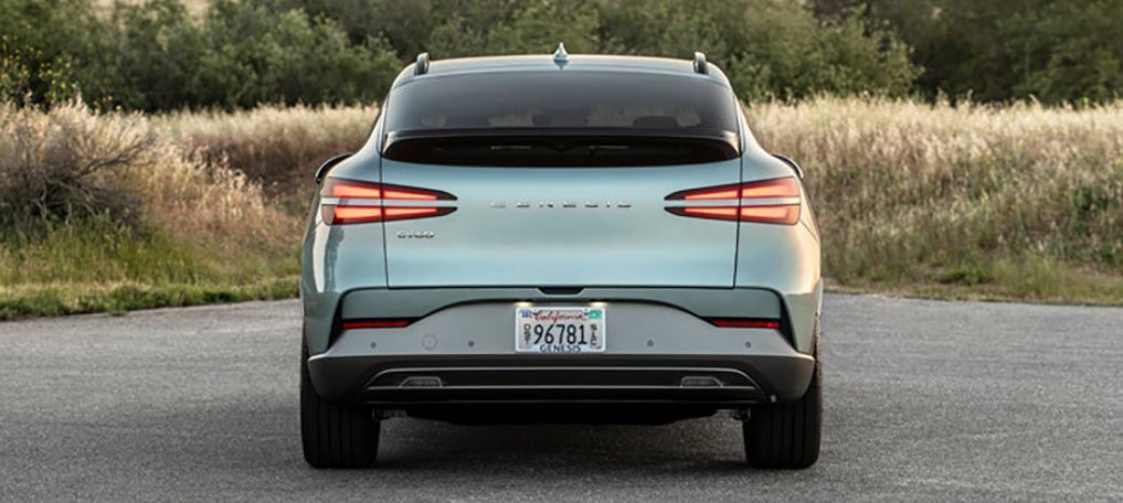 A rear view of a sleek, light blue Genesis GV60 parked in a field with a blurred background of trees and tall grass.