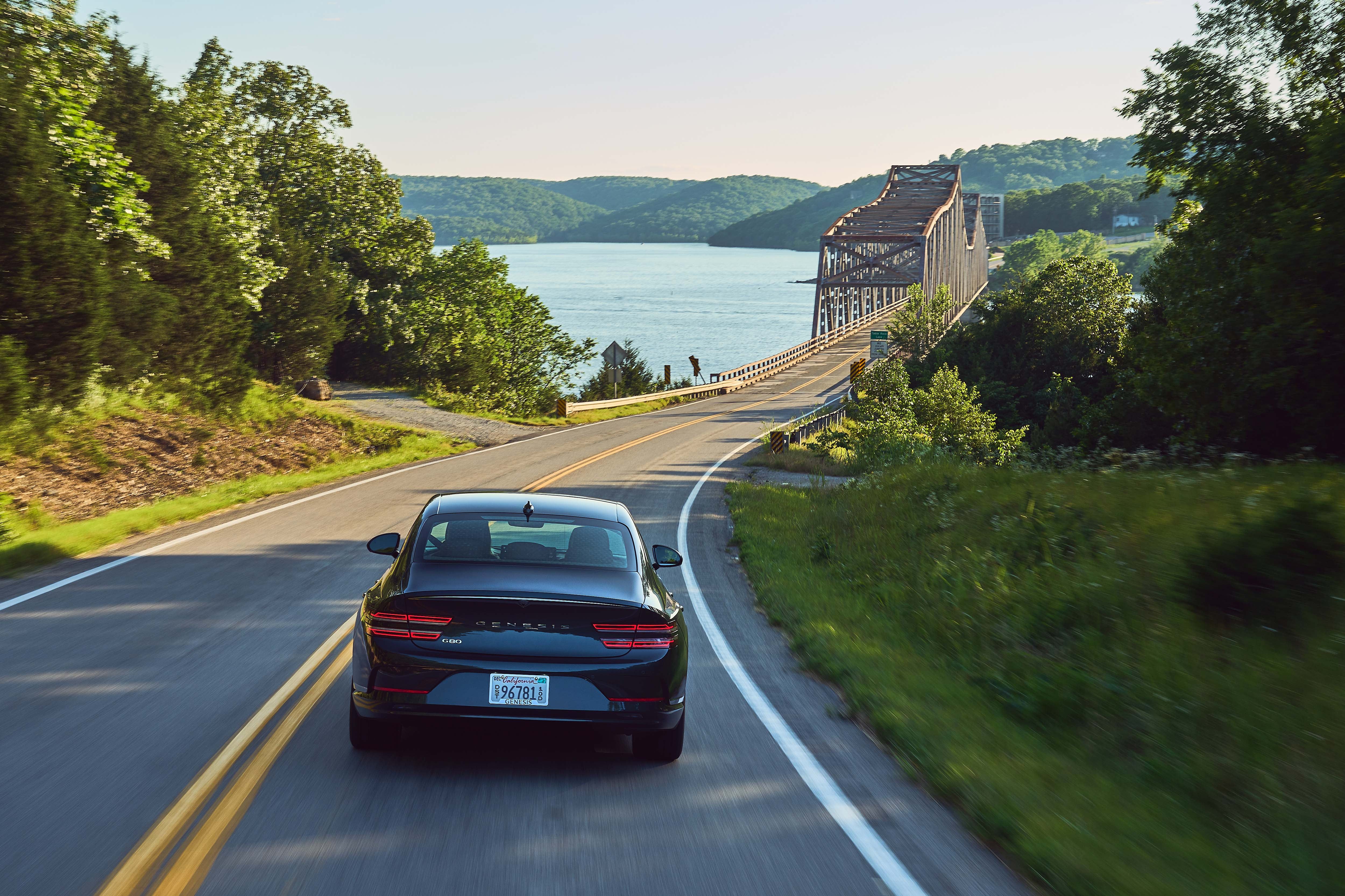 A low, rear view of a black Genesis G80 sedan driving on a winding road with a bridge and a body of water visible in the background. The car is centered on the two-lane road, surrounded by green trees and foliage.