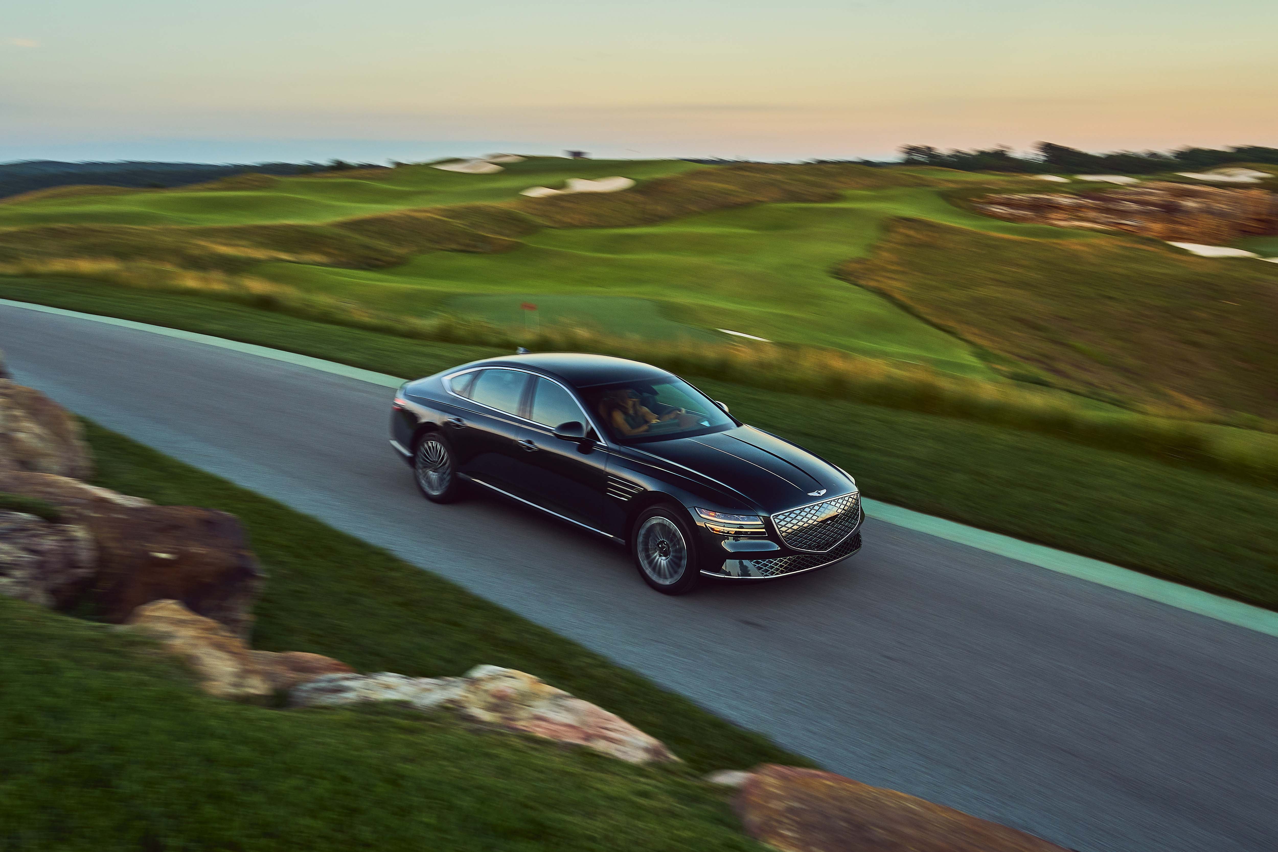 A black Genesis G80 sedan is seen from an aerial view, driving along a paved road that winds through a lush green golf course at sunset.