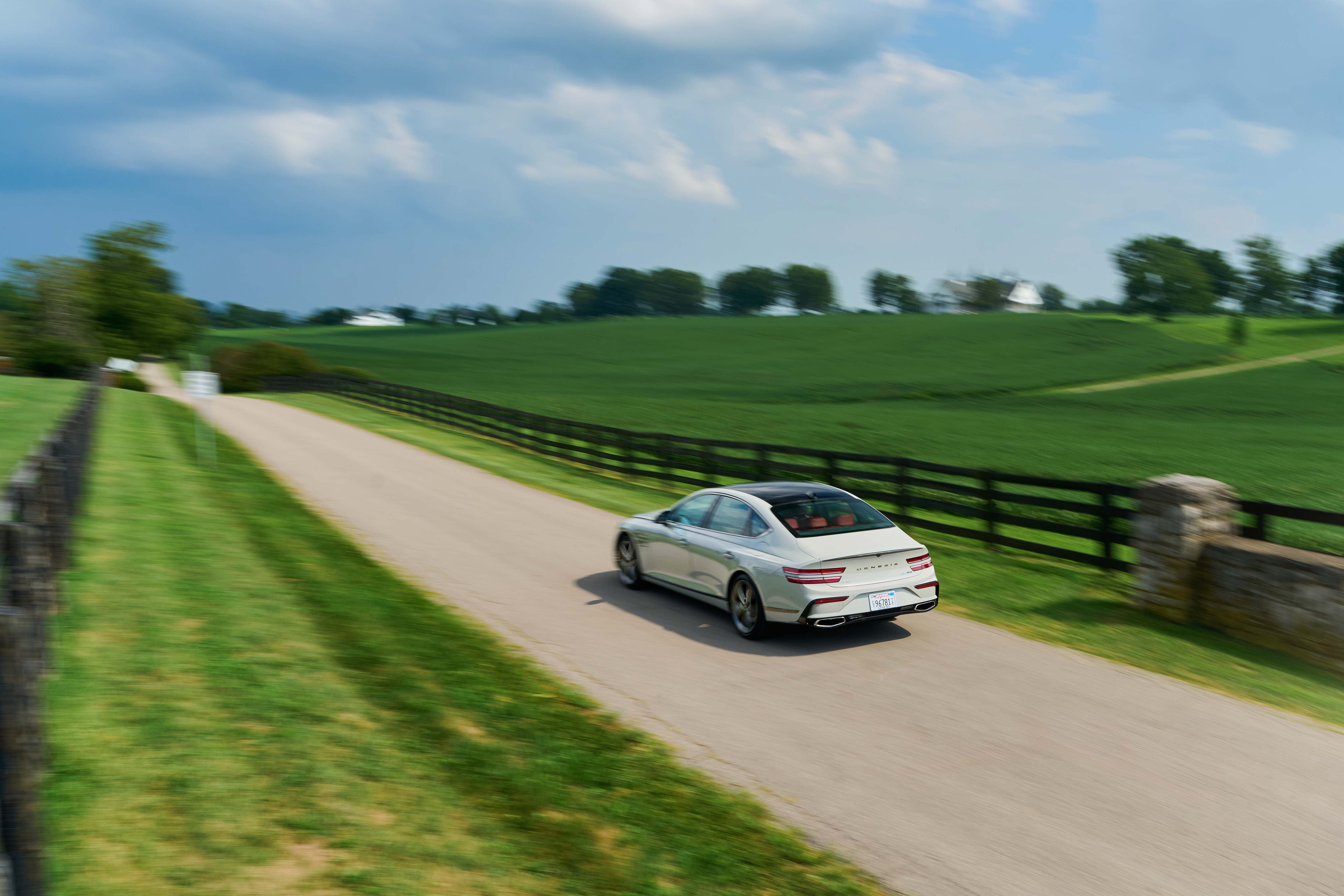 Rear view of a white sedan driving on a paved road through a green, rolling landscape with trees and fields on either side.