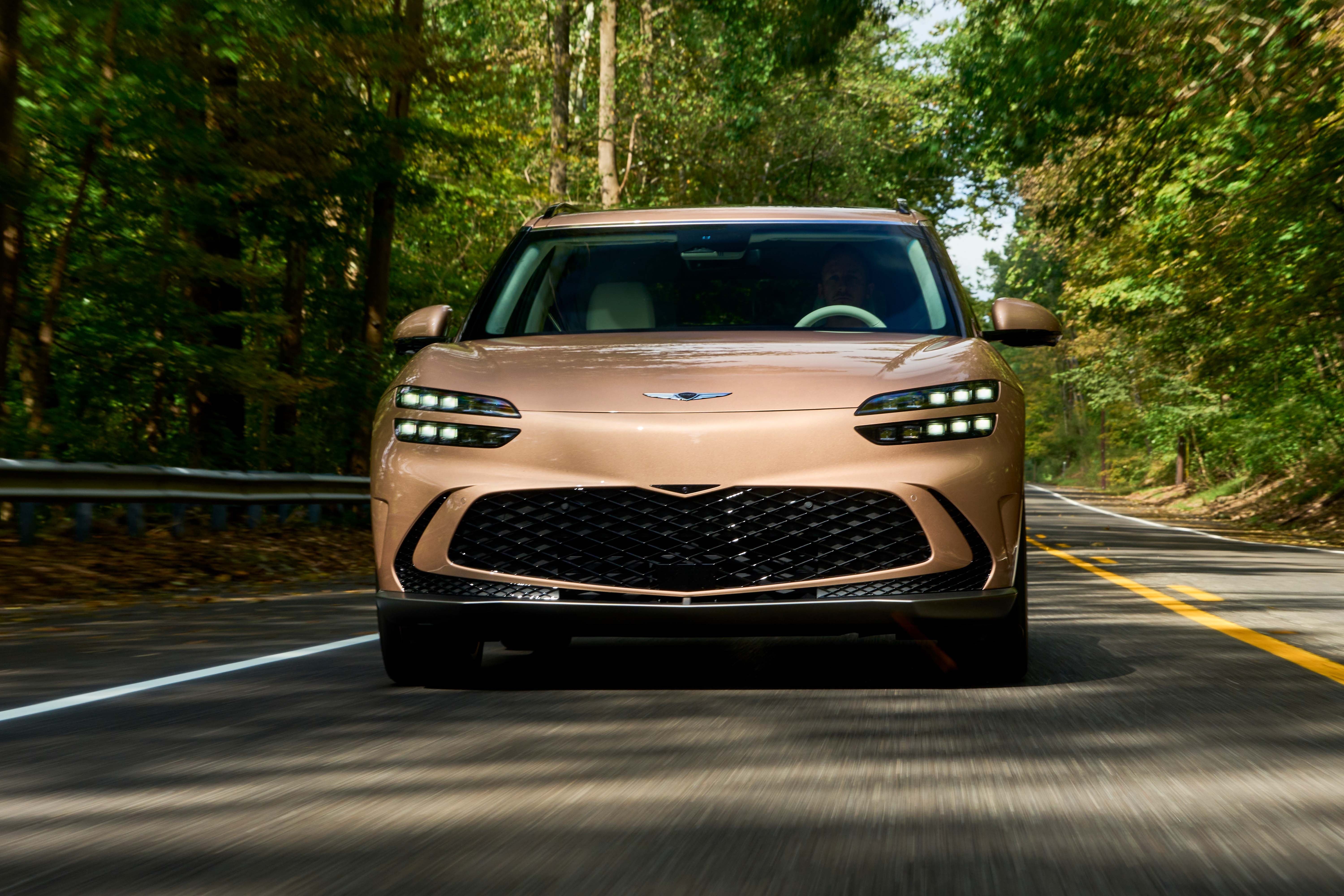 Front view of a bronze Genesis GV70 electric SUV driving on a tree-lined road with sunlight filtering through the trees.