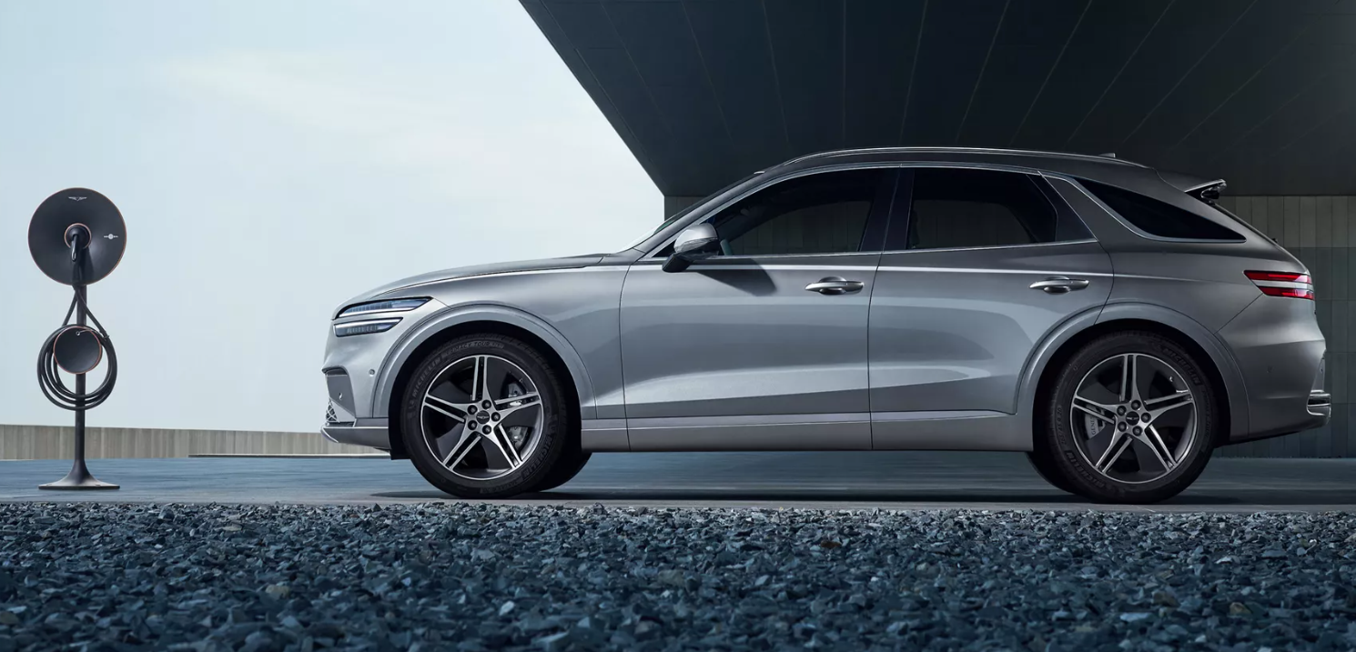 A sleek, silver Genesis GV70 SUV is parked on a dark, pebbled surface next to a modern, dark-colored electric vehicle charging station. The car is viewed from the side, with its elegant lines and intricate wheel design prominent. The background features a minimalist building with large, grey architectural elements, under a bright sky.