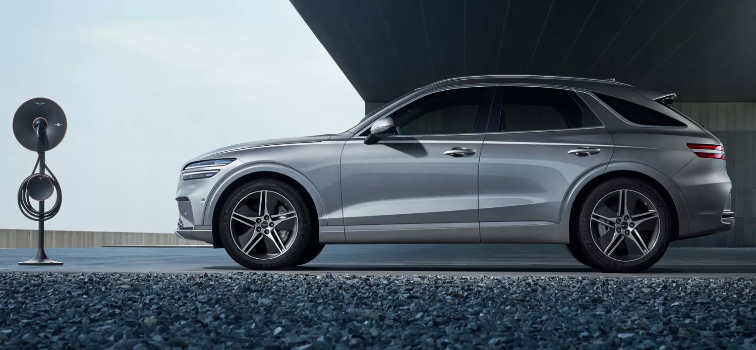 Silver electric SUV parked beside a modern charging station on a gravel surface, with a sleek architectural structure in the background