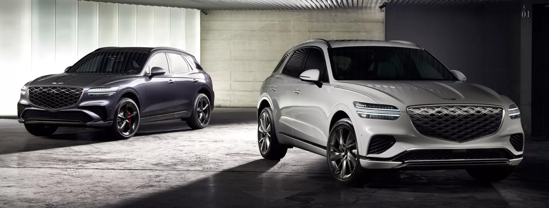 Two Genesis GV70 SUVs are parked in what appears to be a modern, concrete garage or studio with overhead lighting. The SUV on the left is a dark gray or charcoal color, while the one on the right is a light silver or white. Both cars are facing towards the viewer at an angle, showcasing their front grilles, distinct split headlights, and sleek designs.