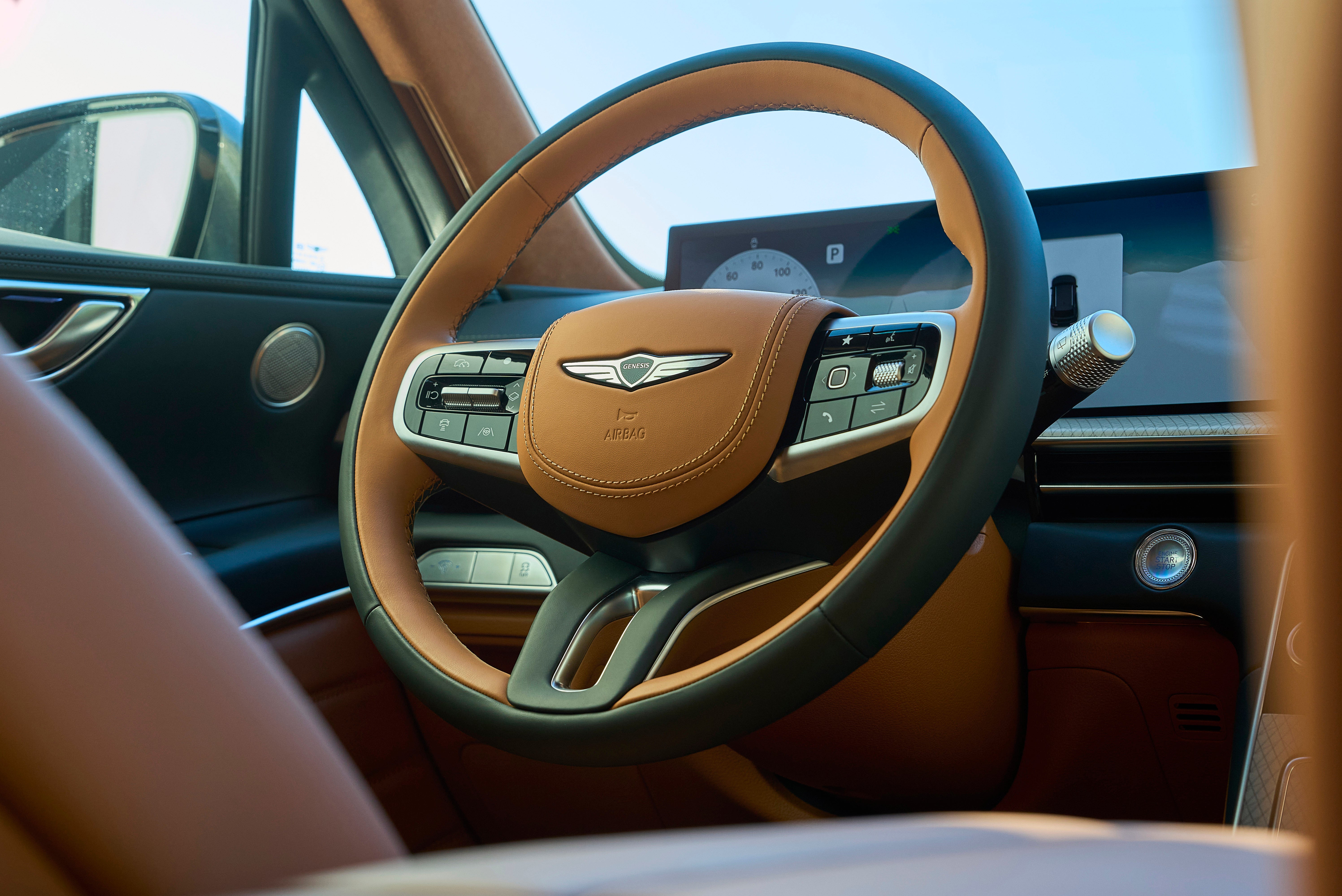Close-up view of the Genesis GV70's tan leather-wrapped steering wheel with integrated controls, digital instrument cluster, and modern interior design elements.