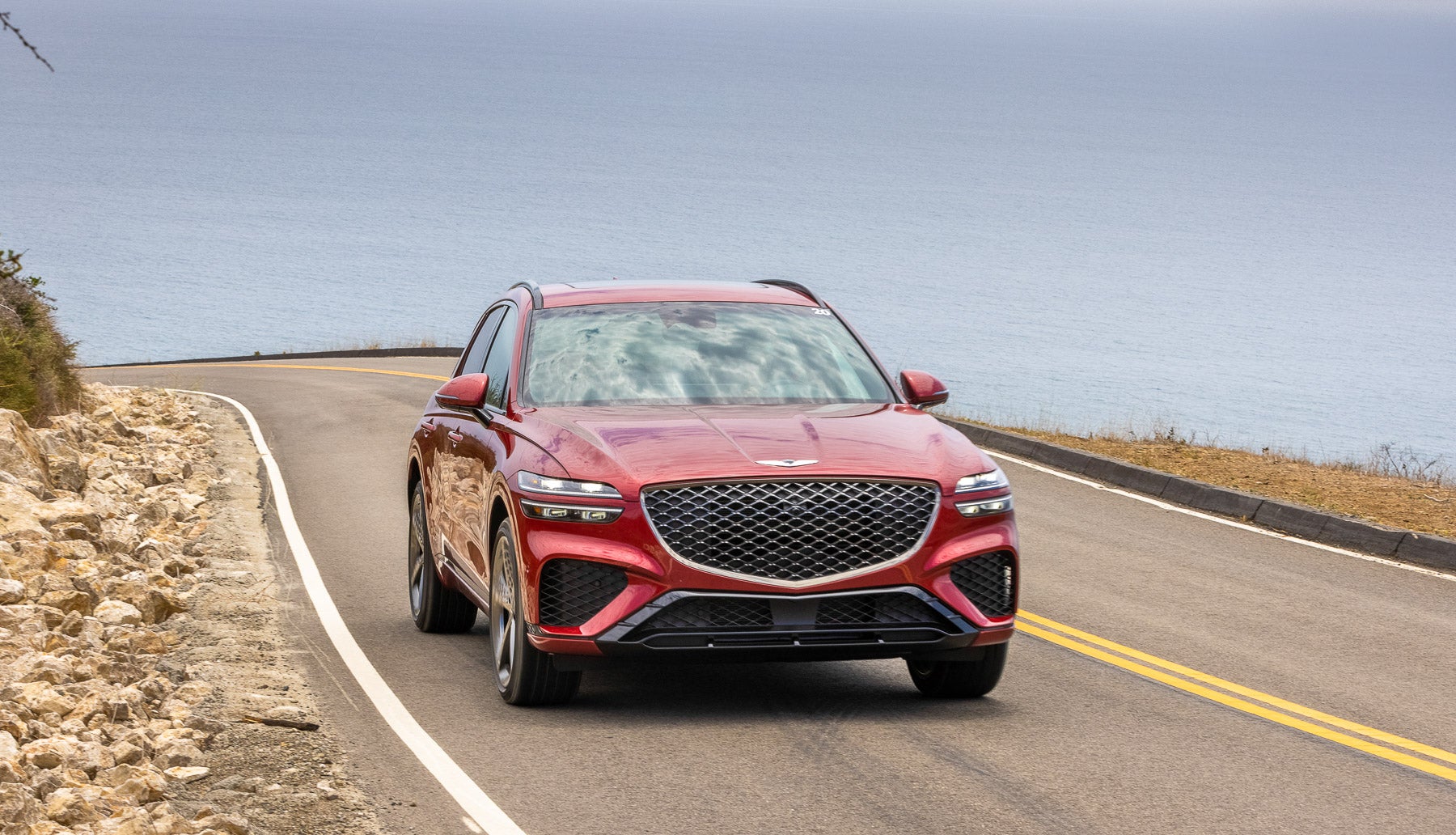 A front view of a bold red Genesis GV80 SUV driving along a scenic coastal road with a beautiful ocean view.