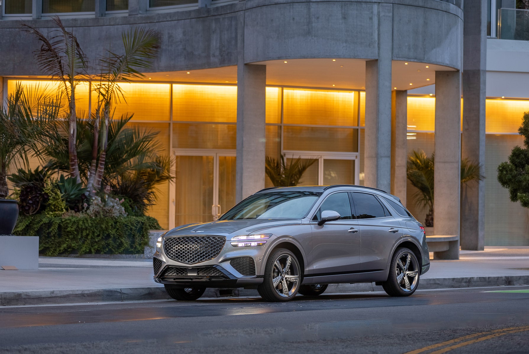 A sleek, silver Genesis GV70 SUV parked in front of a modern building with warm lighting.