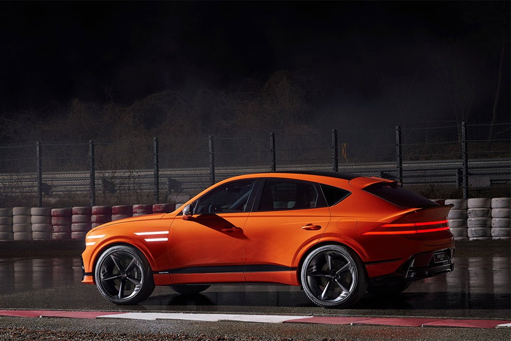 A side view of a sleek, vibrant orange Genesis X Speedium Coupe concept car with dramatic black accents, parked on a wet racetrack under a dark sky.