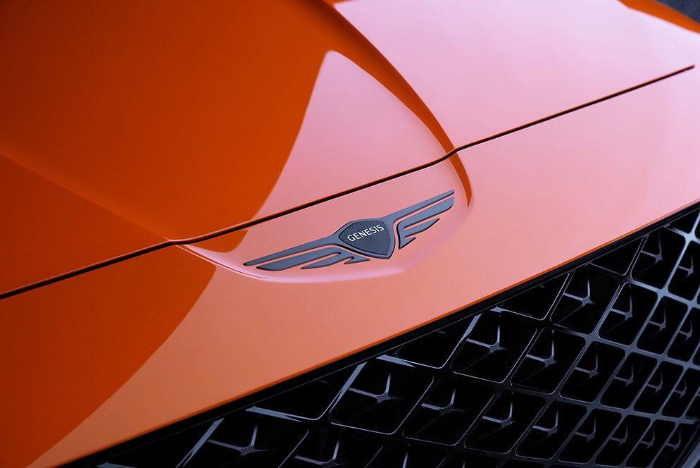 A close-up of the Genesis logo on the hood of a car with a vibrant orange paint finish.