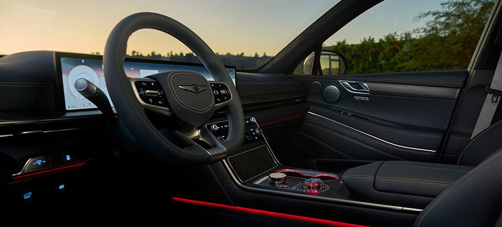 A close-up of the driver's side interior of a Genesis GV80, showcasing the leather seats, steering wheel, dashboard, and ambient lighting.