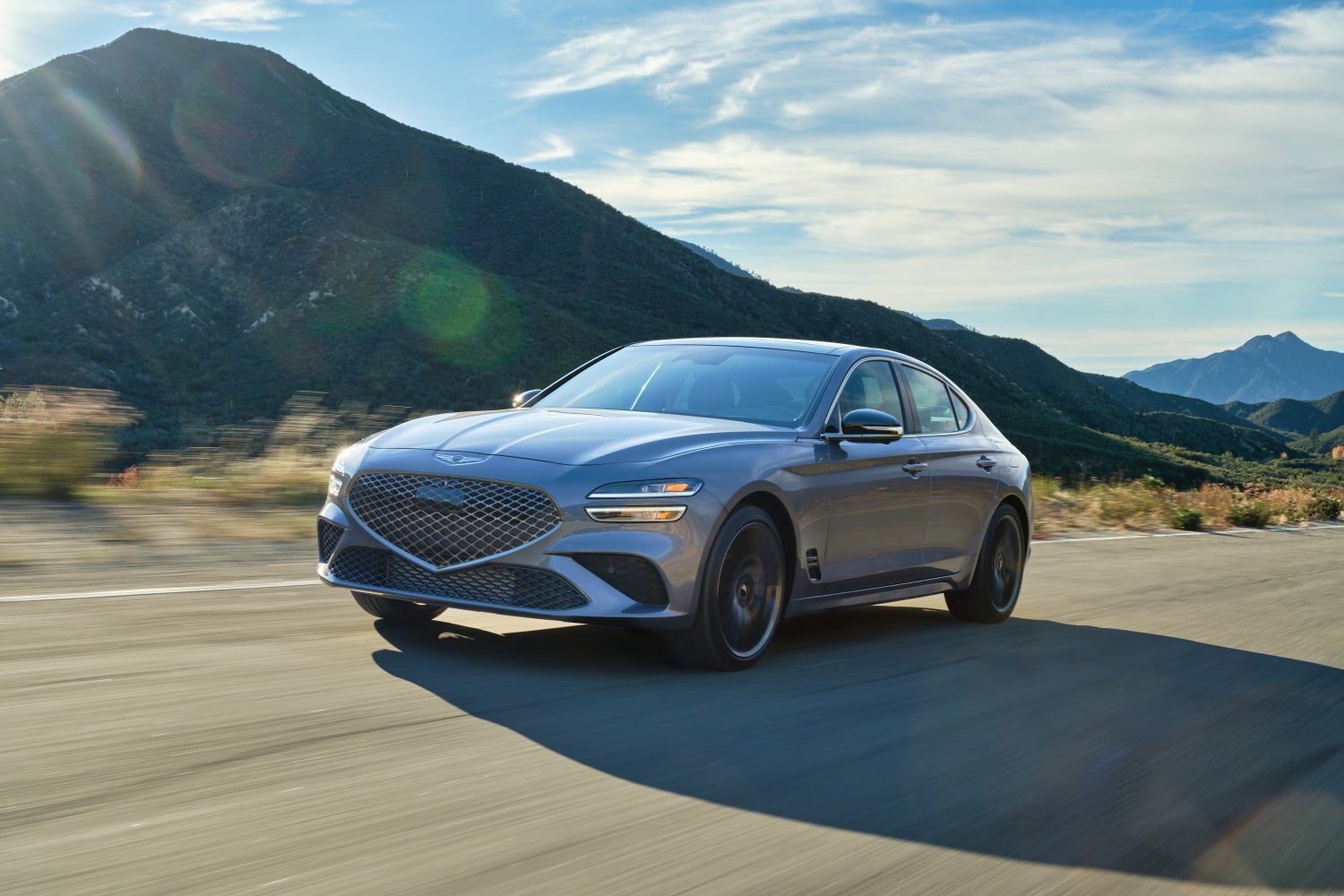 The savile silver 2026 Genesis G70 drives toward the viewer to the left on a rural road in front of a mountainous landscape on a sunny day.