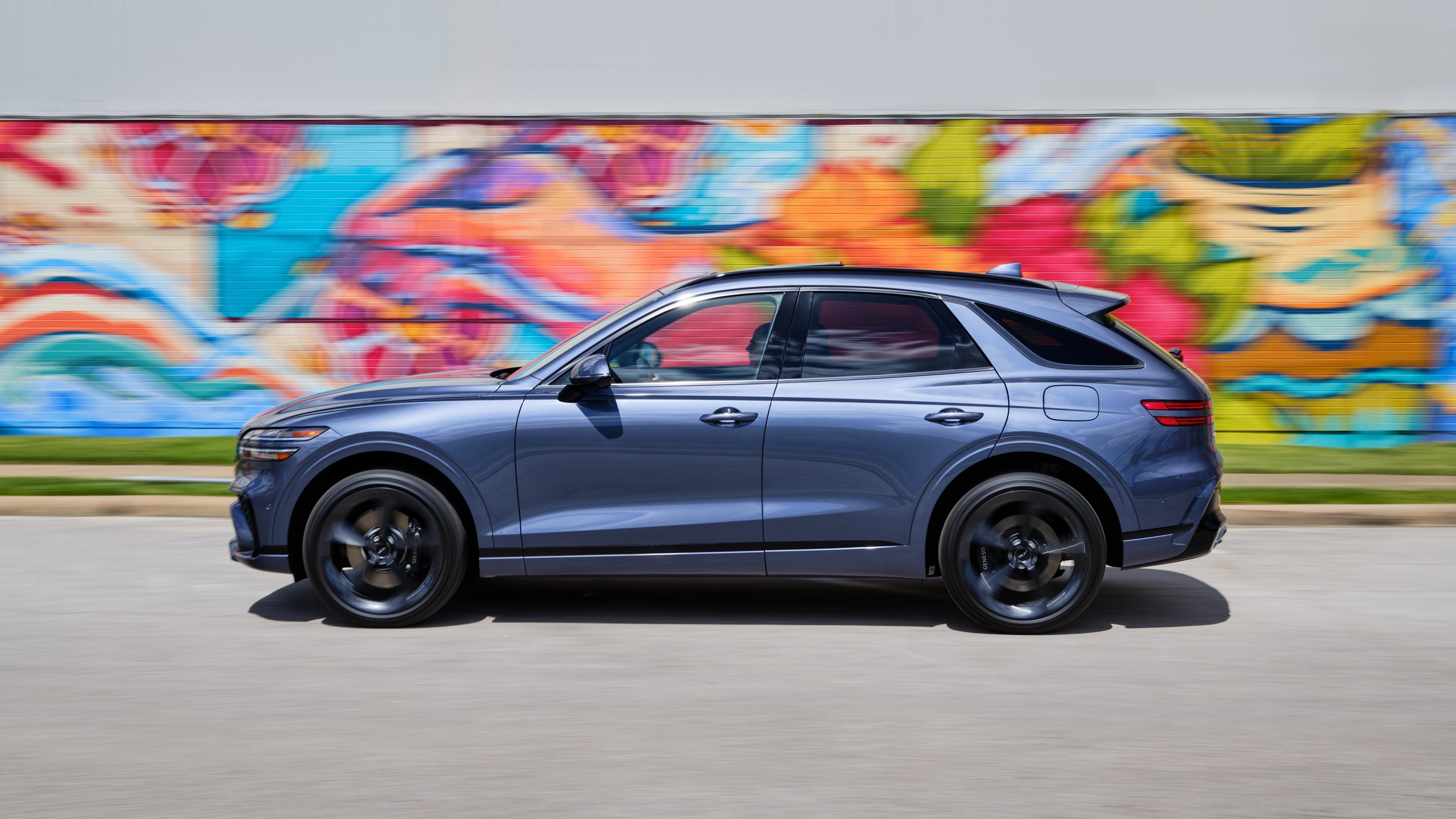 A new GV70 drives directly to the left in front of a wall of colorful graffiti.