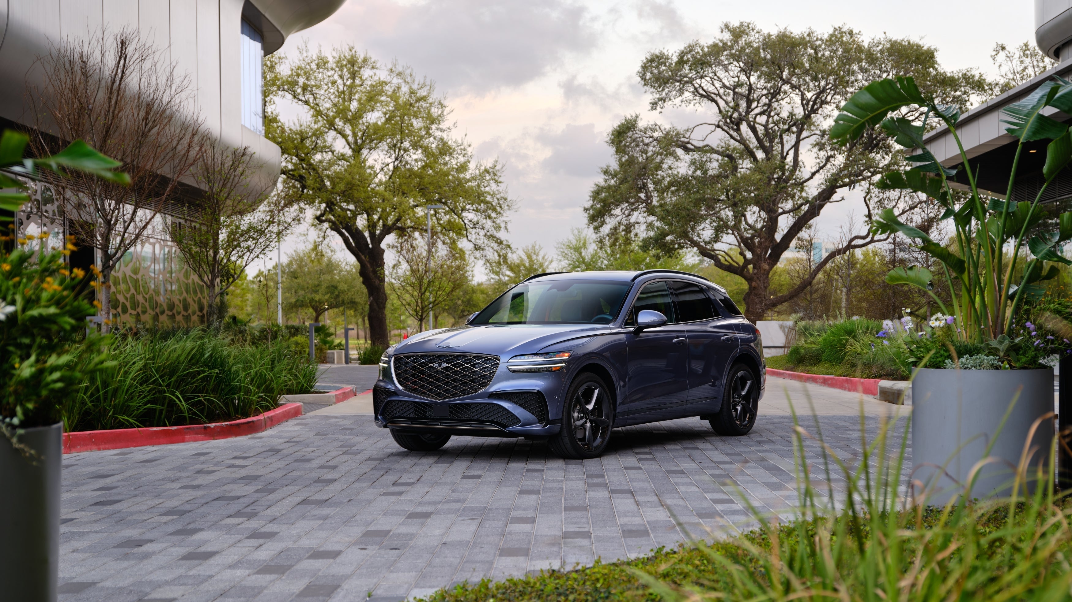 The new Genesis GV70 is parked angled toward the viewer to the left on a tiled driveway surrounded by bright green plantings and the facade of a futuristic building on the far left.