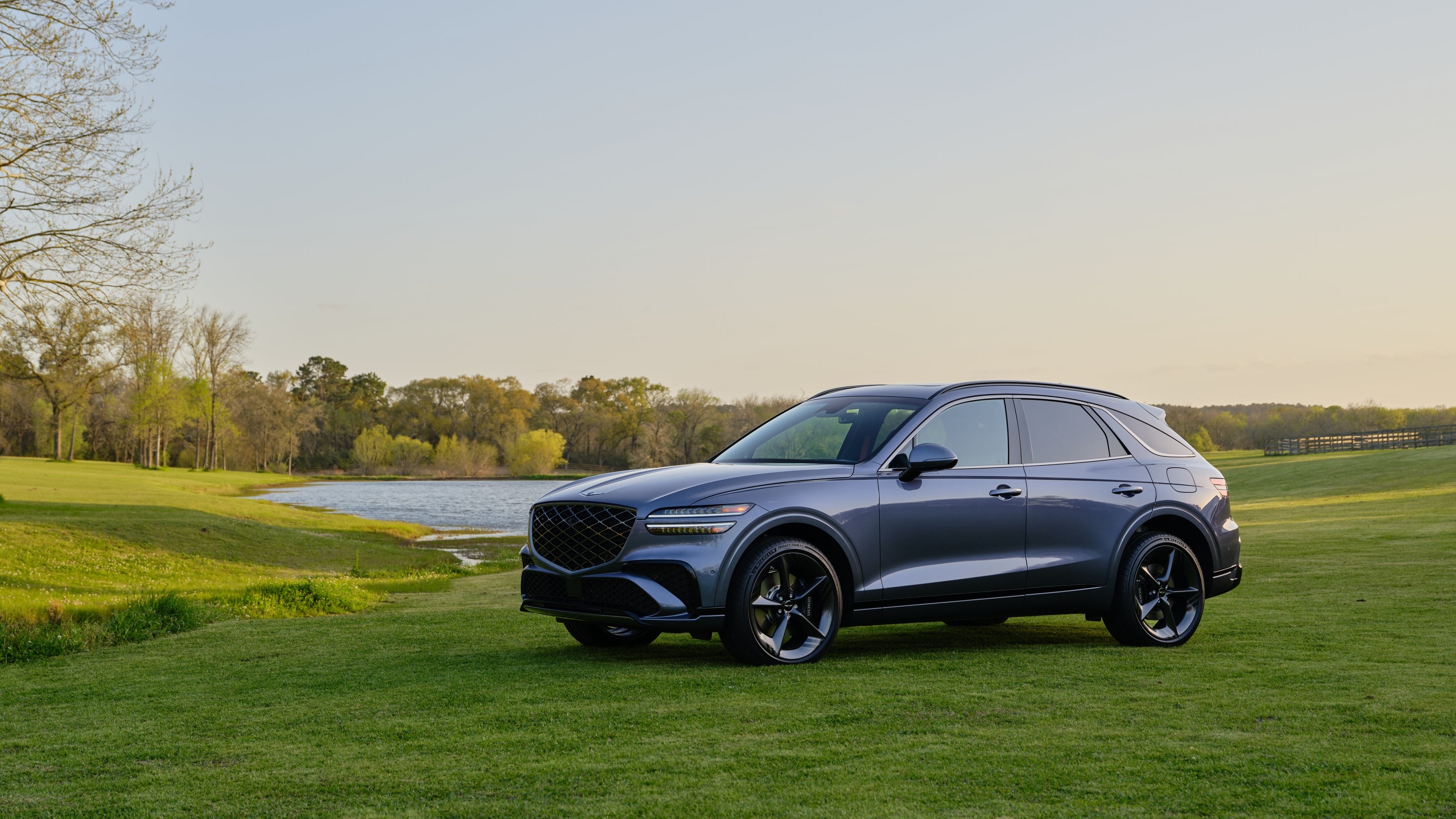 The 2026 Genesis GV70 is parked, angled toward the viewer to the left, on a small bright green grassy hill next to a serene pond.