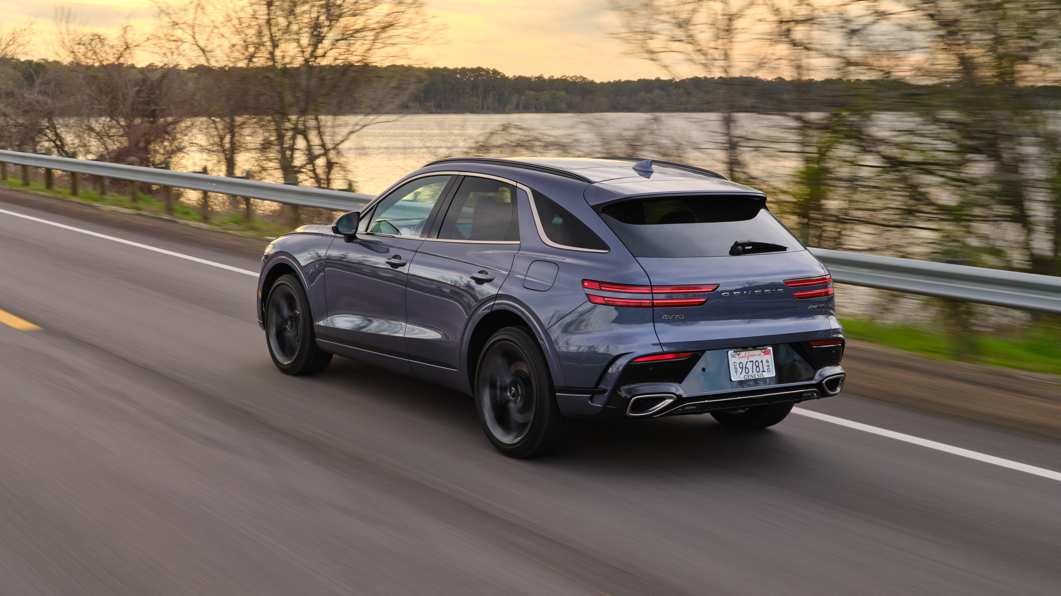 A 2026 GV70 drives away from the viewer to the left on a country road by a lake, seen from a high angle.