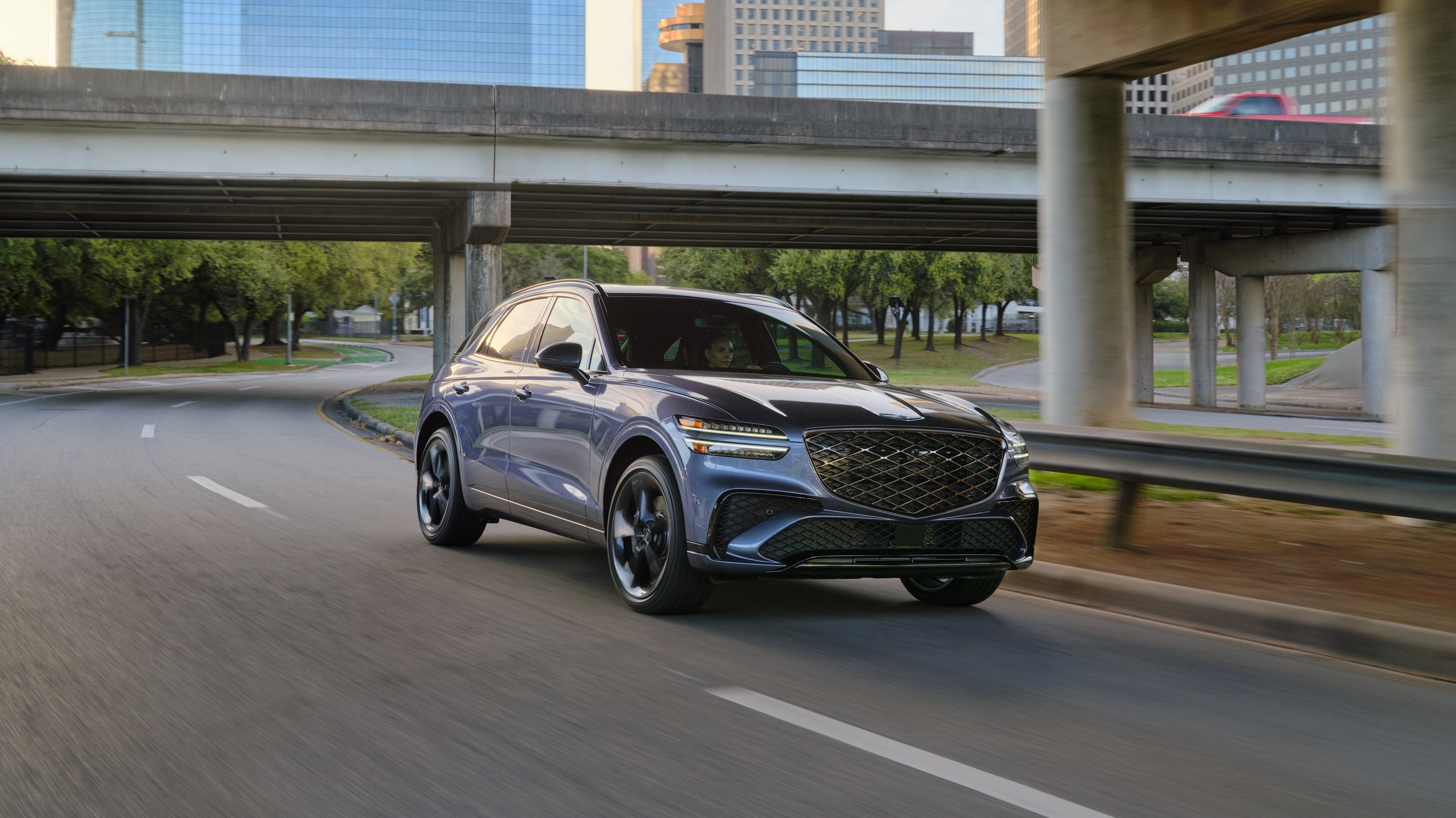 The new GV70 in Chandler drives toward the viewer to the right on a winding city road under an overpass.