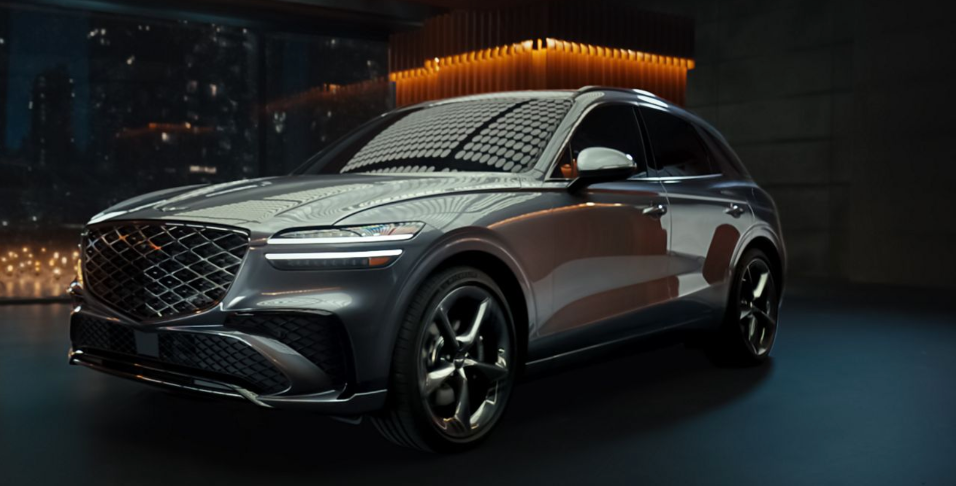 A front three-quarter view of a dark grey Genesis GV70 SUV, showcasing its large, diamond-patterned grille and thin, horizontal headlights. The vehicle is parked indoors in a dimly lit, modern studio.
