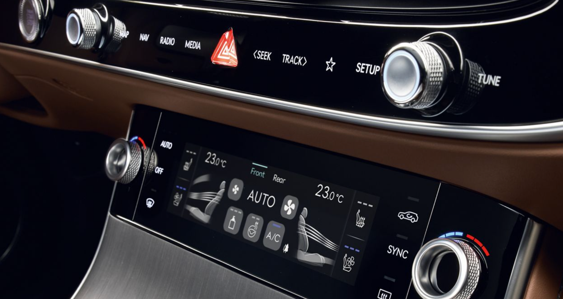 A close-up of the center console of a Genesis GV70 SUV, showing the climate controls, infotainment buttons, and two large, metallic rotary dials. The dashboard is a mix of brown leather, black glossy plastic, and silver trim.