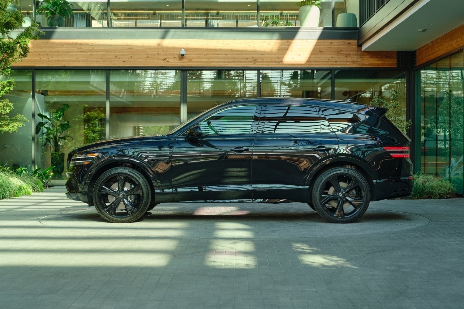 A new vik black Genesis GV80 is parked facing left in front of the glass front of a building with plants all around.