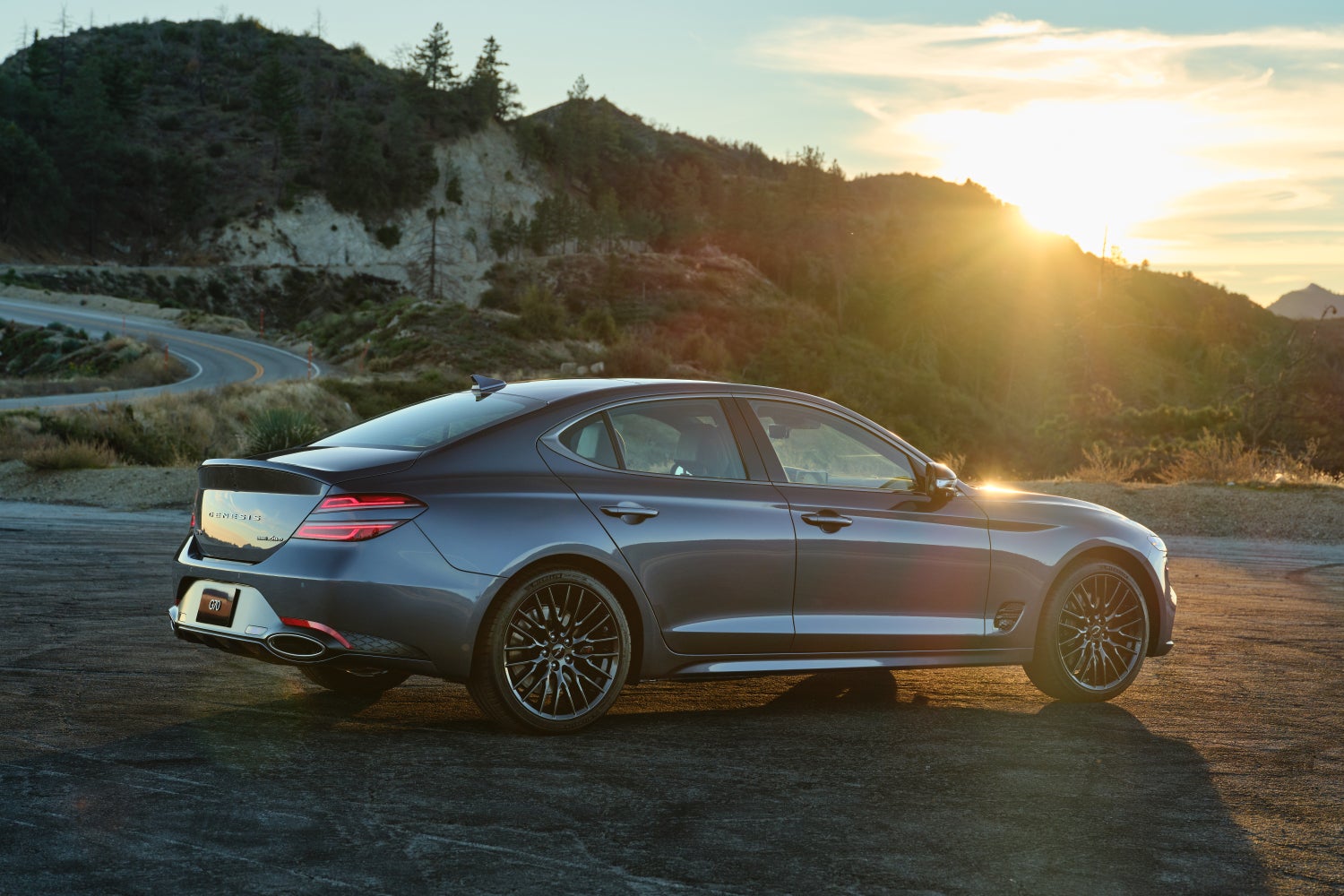 The Prestige Graphite 2026 Genesis G70 in Chandler is parked facing away from the viewer to the right on a gravel turnoff beside a windy mountain road at sunset.