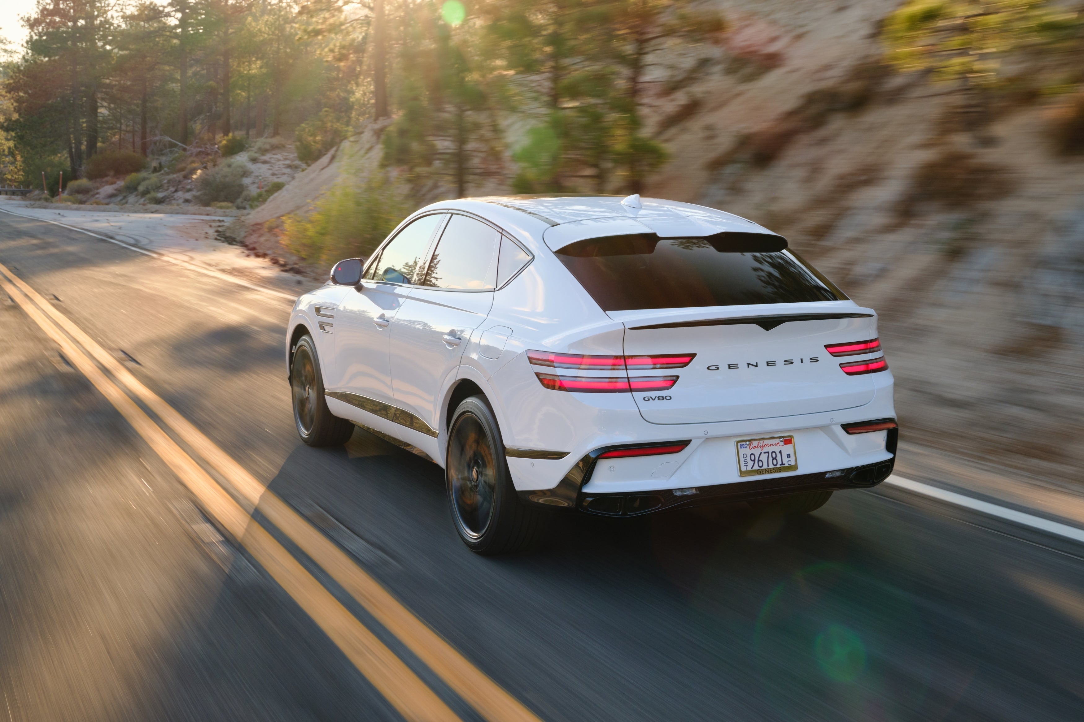 The Genesis GV80 Coupe is driving away from the viewer in Chandler to the left on a mountain road through young evergreen trees.