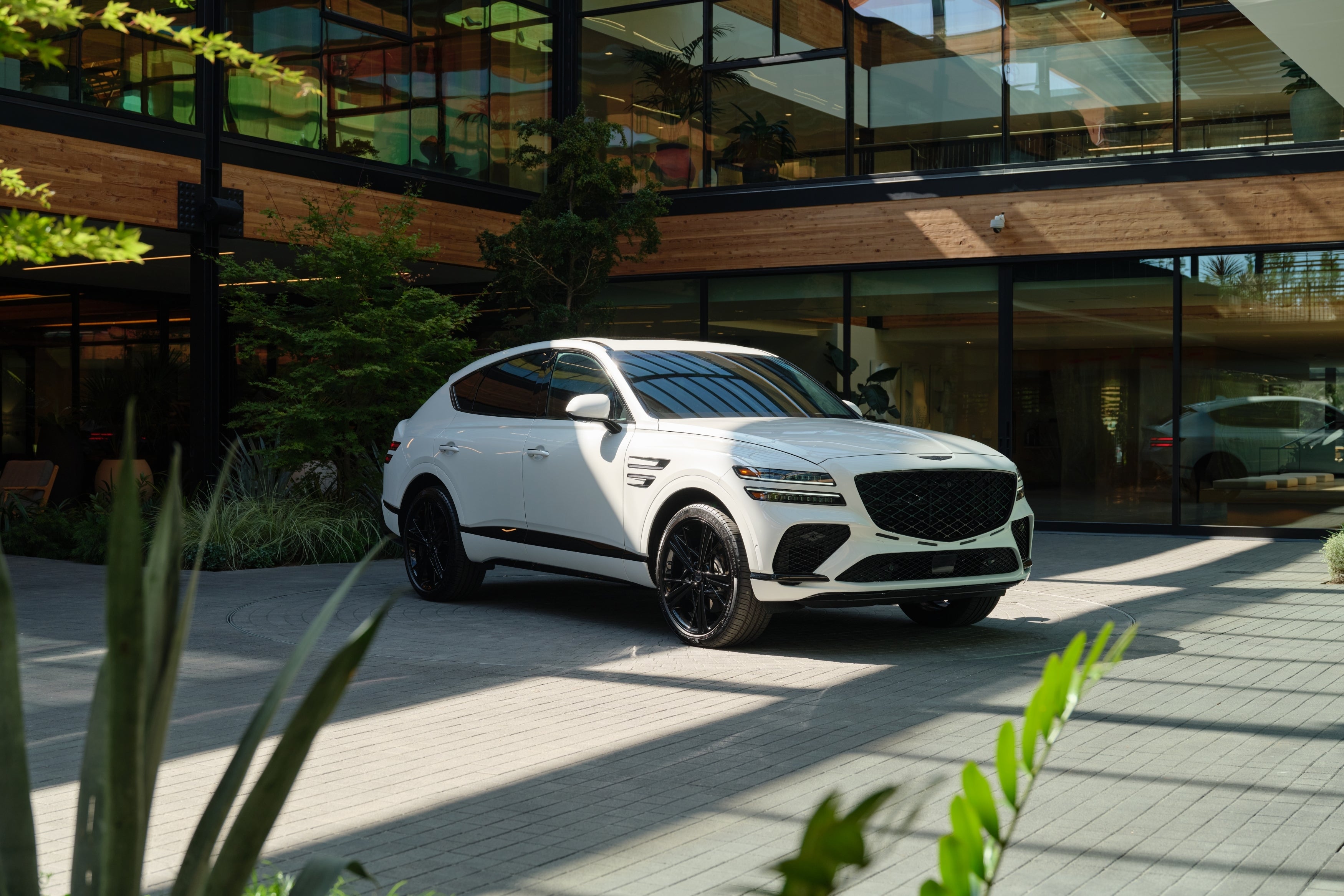 The uyuni white new Genesis GV80 Coupe is parked facing the viewer to the right in front of a modern building made of wood and glass and surrounded by plants.