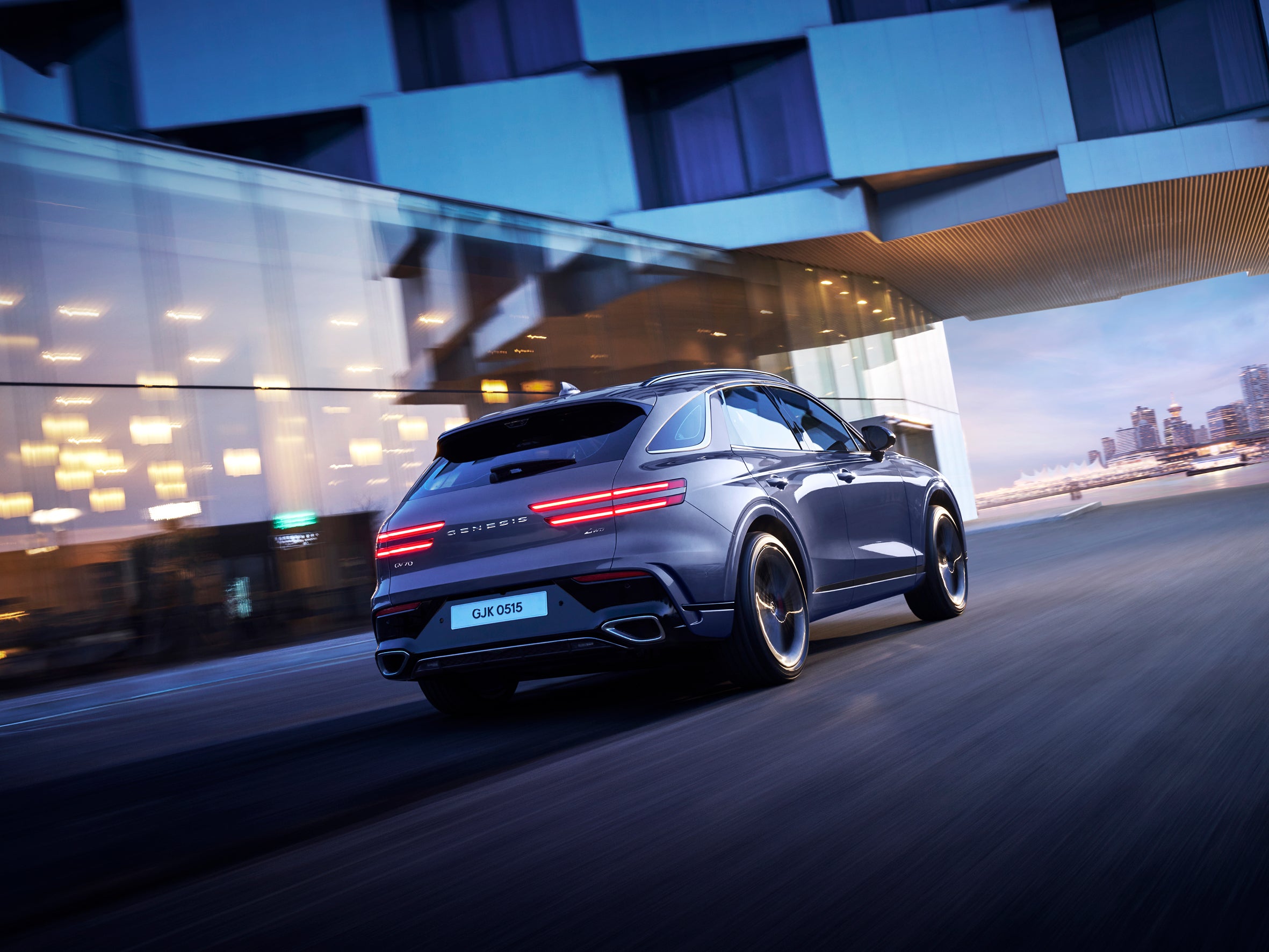 A ceres blue new eGV70 drives away from the viewer to the right in a city at dusk next to a futuristic glass building and overhead bridge. The image is tilted for a distorted perspective of the Genesis electric SUV in Phoenix.