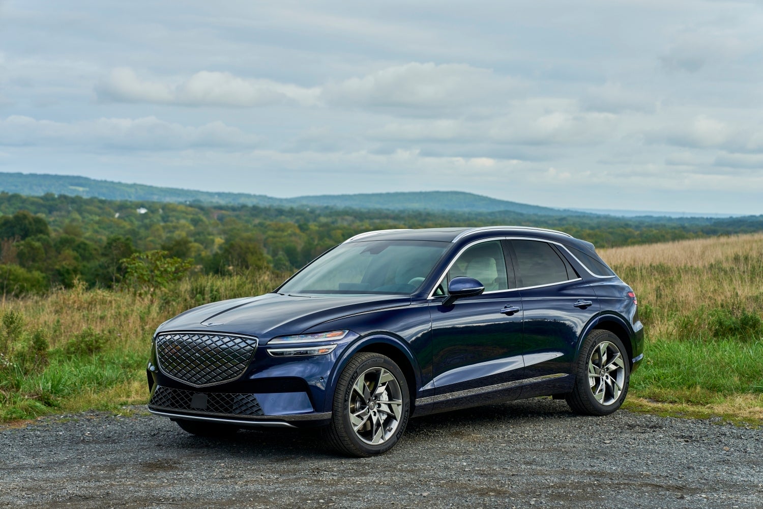 A ceres blue Genesis eGV0 is parked toward the viewer angled left on a gravel road in front of a wild grass field and rolling hills in the background.