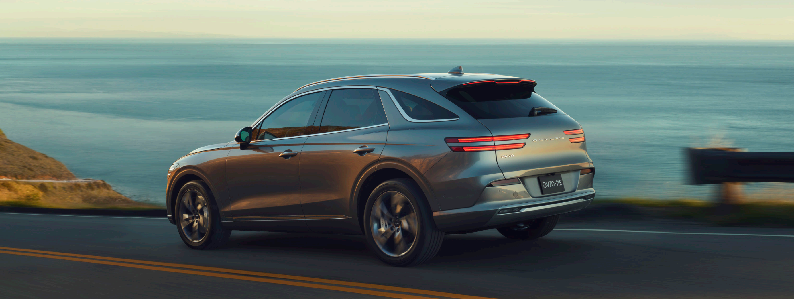 A rear view of a sleek, silver Genesis GV60 driving along a coastal road with a beautiful ocean view and a dramatic sunset in the background.