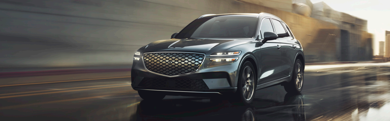A front view of a sleek, dark gray Genesis GV80 driving through a city street in the early morning light.