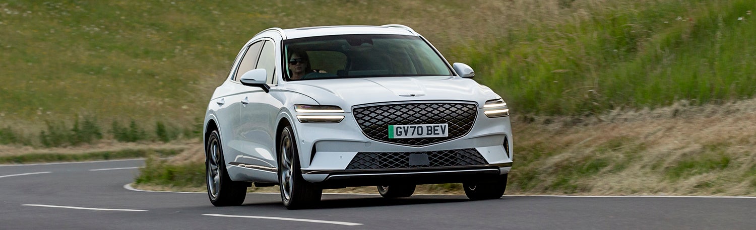 A front view of a sleek, white Genesis GV70 EV driving along a winding road with a lush green field in the background.