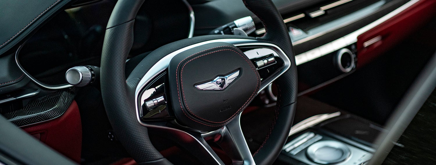 A close-up of a leather-wrapped steering wheel with the Genesis logo and various controls.