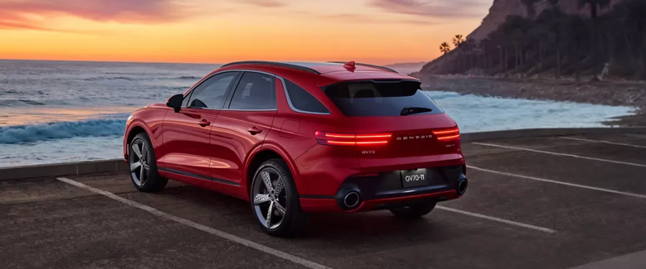 A rear view of a sleek, red Genesis GV70 parked in a coastal parking lot with a stunning sunset over the ocean in the background.